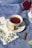 A white teacup filled with tea is placed on a saucer, with a slice of lemon and a cinnamon stick nearby. The setup is on a decorative blue tablecloth with floral patterns. A floral-patterned napkin is folded next to the cup. Red and pink roses in a white vase are partially visible on the right.