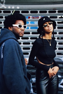 Two individuals are standing against a metal shutter. The person on the left is wearing a dark blue hooded sweatshirt and white sunglasses. The person on the right is wearing a black top with cutouts and black leather pants, accessorized with a black bucket hat and sunglasses.