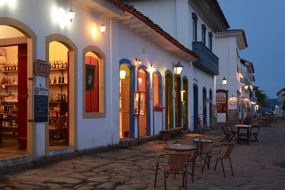 A cobblestone street in Paraty at dusk, with lit shopfronts and café tables