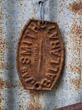 A weathered, rusted metal tag with embossed lettering, attached to a corrugated metal surface. The tag features the words 'JAS SMITH BALLARAT.' The metal surface also appears rusted, adding texture and character to the scene.