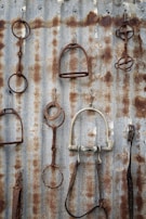 A variety of rusted horse tack, including stirrups and bits, hang on a corrugated metal wall. Each piece displays varying degrees of rust and wear, contrasting with the metallic background. The arrangement creates a rustic and vintage aesthetic.