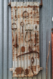 A collection of rusty, metal objects is arranged on a piece of corrugated metal. The objects include various hooks, rings, and implements, showcasing a weathered and aged appearance. The backdrop is a corrugated metal surface, adding to the industrial aesthetic.