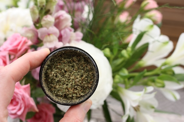 A hand holding a small container filled with a green, dried herbal mixture against a backdrop of assorted blooming flowers including pink, white, and light purple blossoms.
