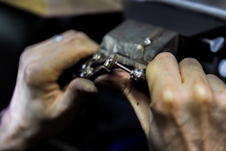 Hands engaged in a detailed task involving a small mechanical device, possibly a watch or some type of precision instrument. The workspace appears to be dimly lit, focusing on the fine manual work being executed.