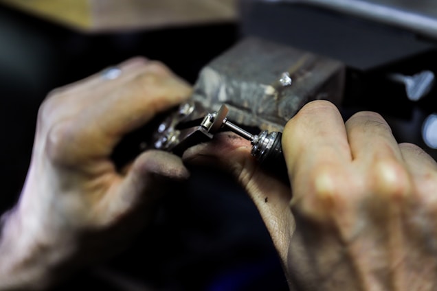 Hands engaged in a detailed task involving a small mechanical device, possibly a watch or some type of precision instrument. The workspace appears to be dimly lit, focusing on the fine manual work being executed.