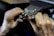 Hands engaged in a detailed task involving a small mechanical device, possibly a watch or some type of precision instrument. The workspace appears to be dimly lit, focusing on the fine manual work being executed.