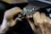 Hands engaged in a detailed task involving a small mechanical device, possibly a watch or some type of precision instrument. The workspace appears to be dimly lit, focusing on the fine manual work being executed.