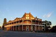 A historic two-story building with ornate architectural features, including arched windows and decorative railings. The structure appears well-preserved with a red and white color scheme. Trees flank the building and the sky is clear with soft clouds in the background.
