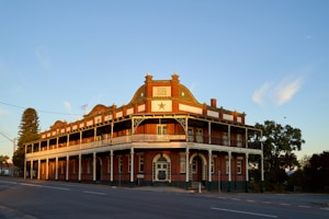A historic two-story building with ornate architectural features, including arched windows and decorative railings. The structure appears well-preserved with a red and white color scheme. Trees flank the building and the sky is clear with soft clouds in the background.
