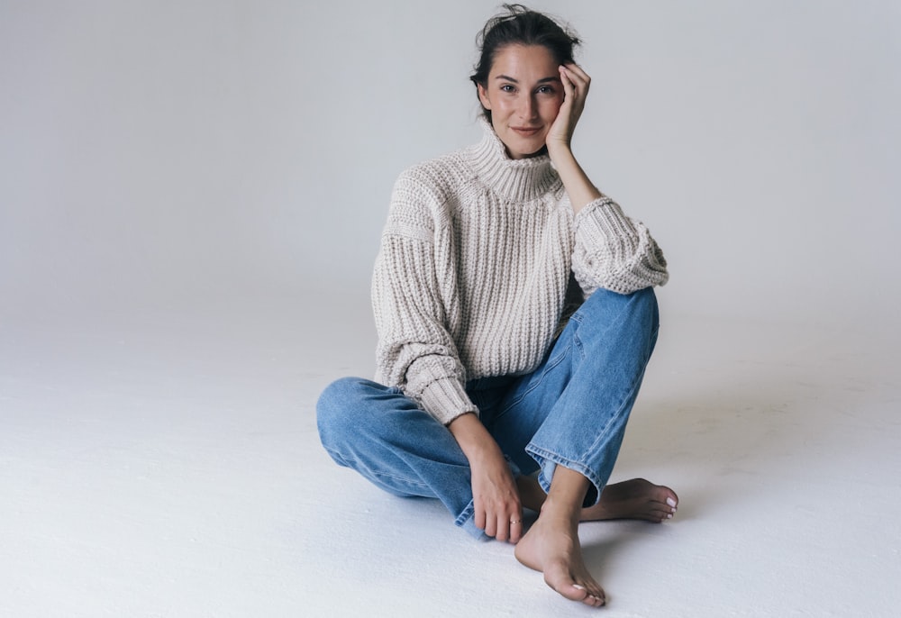 Femme en pull gris et jean bleu assis sur un sol blanc