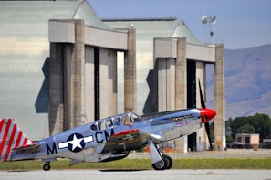 A vintage silver aircraft with red accents parked on a tarmac in front of large hangars. The aircraft features a distinctive World War II star insignia and red-striped tail. The background includes expansive hangar doors and a clear, blue sky.