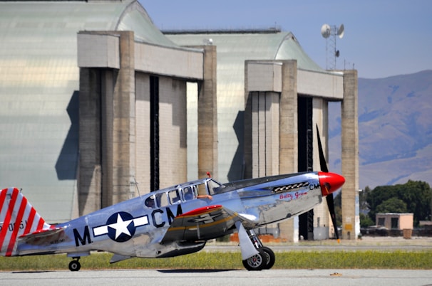 A vintage silver aircraft with red accents parked on a tarmac in front of large hangars. The aircraft features a distinctive World War II star insignia and red-striped tail. The background includes expansive hangar doors and a clear, blue sky.