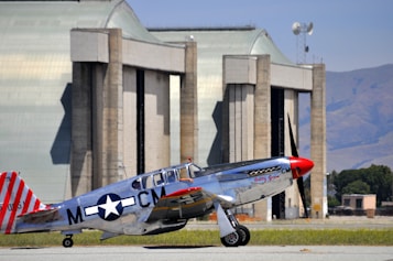 A vintage silver aircraft with red accents parked on a tarmac in front of large hangars. The aircraft features a distinctive World War II star insignia and red-striped tail. The background includes expansive hangar doors and a clear, blue sky.