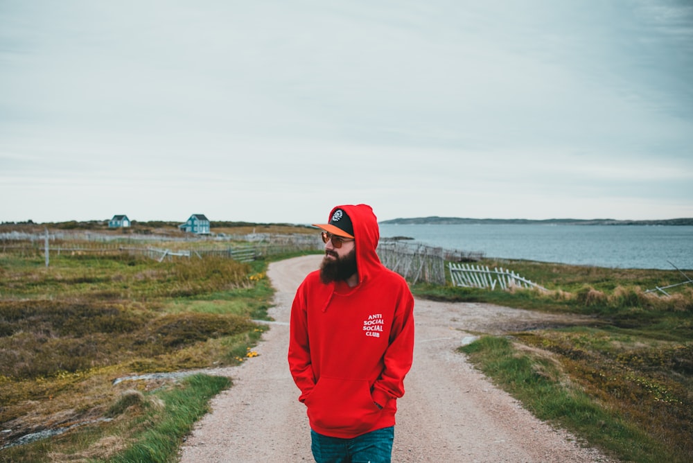 man in red hoodie standing on gray sand near body of water during daytime