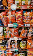 A variety of snack bags displayed on store shelves, including brands like Doritos, Ruffles, and Lay's. The packaging is colorful, featuring reds, blues, greens, and oranges, with visible pricing tags showing 5.50 TL. The bags have promotional stickers attached.