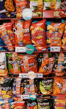 A variety of snack bags displayed on store shelves, including brands like Doritos, Ruffles, and Lay's. The packaging is colorful, featuring reds, blues, greens, and oranges, with visible pricing tags showing 5.50 TL. The bags have promotional stickers attached.