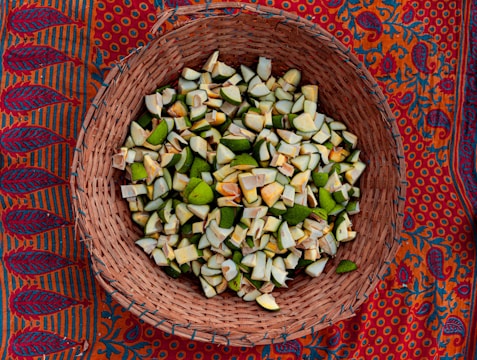A wicker basket filled with chopped, green and white unripe fruit pieces is placed on a brightly colored, intricate patterned fabric featuring red, blue, and orange hues.