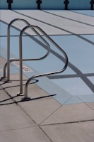 A metal handrail is attached to the concrete deck of an empty swimming pool. The pool's painted surface exposes its lanes, which are marked by black lines on a light blue background. Shadows of the railings cast sharp patterns on the deck.