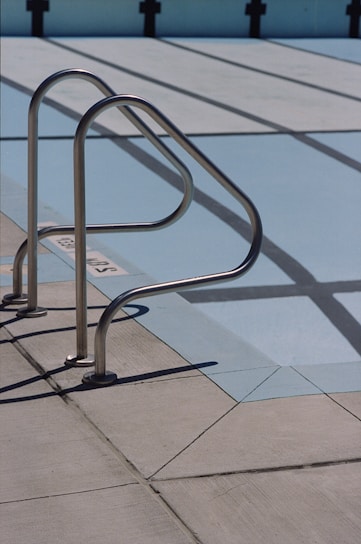 A metal handrail is attached to the concrete deck of an empty swimming pool. The pool's painted surface exposes its lanes, which are marked by black lines on a light blue background. Shadows of the railings cast sharp patterns on the deck.