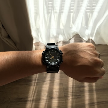A wrist with a watch featuring a black strap and dial is extended in front of a background with white curtains letting in sunlight. The wrist and watch are prominently in focus against a slightly blurred backdrop.