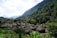 A village with round thatched-roof huts nestled in a lush, green valley surrounded by dense forested hills. The sky is partly cloudy, allowing sunlight to softly illuminate the landscape. The huts are arranged closely together, suggesting a small, tight-knit community within a natural setting.
