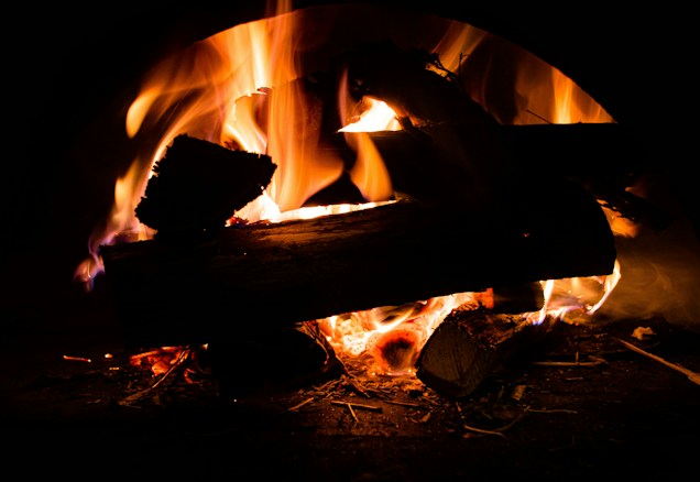 Logs burning with vibrant flames, displaying rich orange and yellow hues with glowing embers visible below. The scene is set in a dark background, enhancing the warmth and intensity of the fire.