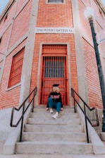 A person is sitting on concrete steps leading to a red door of a brick building. They are wearing a graduation cap and a sweatshirt with the words 'El Paso' printed on it. The building is labeled 'Co-Op Rio Cattle Co.' and features a street lamp to the right.
