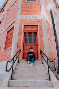 A person is sitting on concrete steps leading to a red door of a brick building. They are wearing a graduation cap and a sweatshirt with the words 'El Paso' printed on it. The building is labeled 'Co-Op Rio Cattle Co.' and features a street lamp to the right.