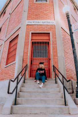 A person is sitting on concrete steps leading to a red door of a brick building. They are wearing a graduation cap and a sweatshirt with the words 'El Paso' printed on it. The building is labeled 'Co-Op Rio Cattle Co.' and features a street lamp to the right.