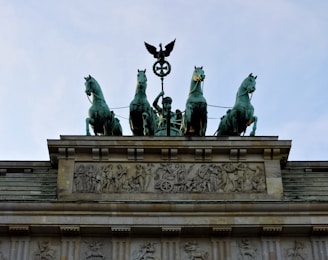 A quadriga statue depicting a chariot pulled by four horses, mounted on top of a historic stone building adorned with intricate carvings and reliefs. The central figure in the chariot holds a staff topped with a circular symbol and an eagle.