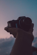 A hand is holding a compact digital camera against a backdrop of a twilight sky. The scene features a mix of warm and cool tones, suggesting a peaceful evening.