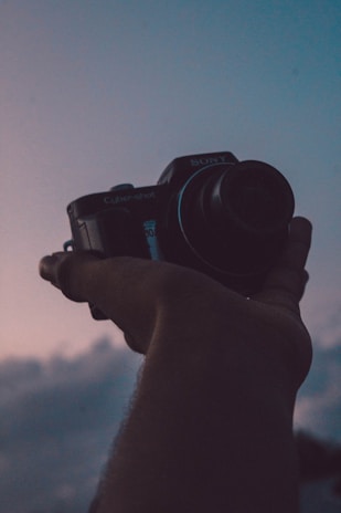 A hand is holding a compact digital camera against a backdrop of a twilight sky. The scene features a mix of warm and cool tones, suggesting a peaceful evening.