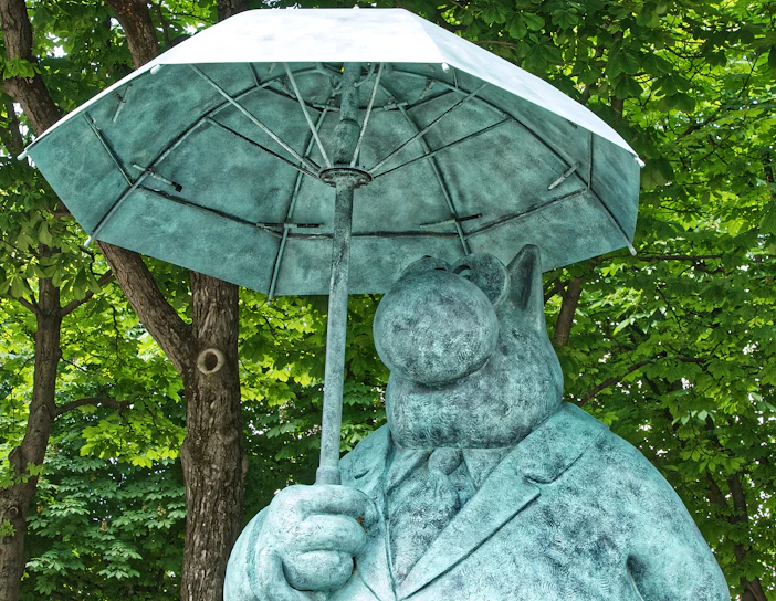 A statue of a cartoonish character resembling an animal is depicted holding an umbrella. The figure is dressed in a formal suit and stands against a backdrop of lush green trees.