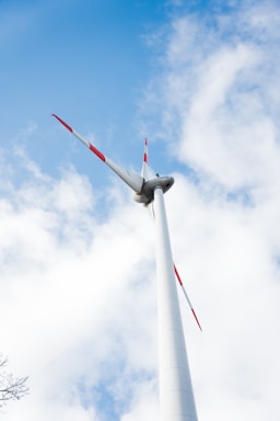 A tall wind turbine with red-tipped blades stands against a backdrop of blue sky and scattered white clouds.