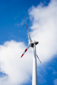 A tall wind turbine with three blades against a backdrop of a clear blue sky and scattered white clouds. The turbine's blades have red tips for visibility.