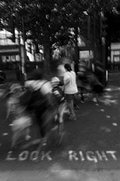 A blurred black and white scene featuring multiple people in motion, possibly crossing a street. A large tree is visible in the background and the words 'LOOK RIGHT' are painted on the pavement.