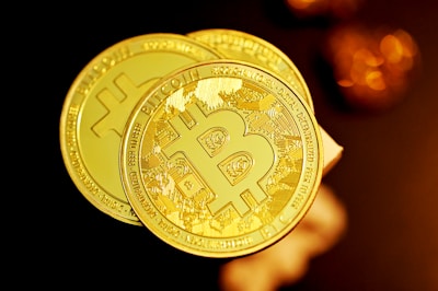 Two shiny gold-colored coins with the Bitcoin emblem are prominently featured, each displaying intricate details and text related to the cryptocurrency. The background has a soft, warm glow with blurred, circular bokeh effects, enhancing the image's focus on the coins.