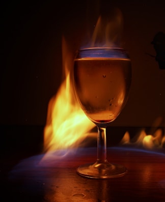 A glass filled with liquid is surrounded by vibrant flames, creating a dramatic and dynamic effect. The flames emit a warm glow, illuminating the glass and casting intricate reflections.