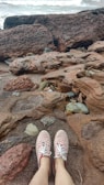 A pair of feet wearing red and white chevron-patterned shoes rest on a rocky beach. The shore is composed of weathered, rugged rocks and stones, with sand visible in between. In the background, waves appear to be crashing gently onto the rocks.