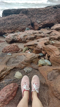 A pair of feet wearing red and white chevron-patterned shoes rest on a rocky beach. The shore is composed of weathered, rugged rocks and stones, with sand visible in between. In the background, waves appear to be crashing gently onto the rocks.