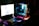 A person is sitting in front of a computer setup with a glowing RGB keyboard and a large monitor displaying a game. The scene is dimly lit, and the person is wearing a headset, suggesting they might be playing video games or engaged in online communication. The internal components of the computer tower are visible and illuminated in blue and green lights.