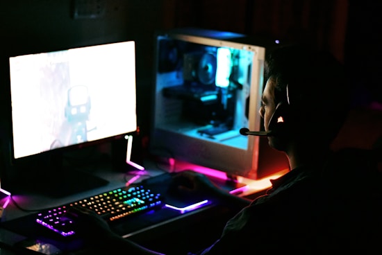 A person is sitting in front of a computer setup with a glowing RGB keyboard and a large monitor displaying a game. The scene is dimly lit, and the person is wearing a headset, suggesting they might be playing video games or engaged in online communication. The internal components of the computer tower are visible and illuminated in blue and green lights.