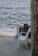 A white wrought iron bench is positioned on a stone terrace overlooking the ocean. Waves gently crash onto the sandy shore. A blackboard leaning against the terrace wall has chalk writings in Italian, likely announcing the availability of dinner and other offerings.
