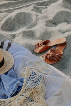 A relaxed beach setting featuring a striped towel laid on the sand, a pair of brown sandals, a straw hat, and a mesh bag with a denim shirt partially visible.