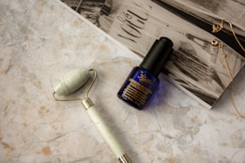 A facial roller made of jade and a small bottle of a skincare product are placed on a marbled surface. The blue bottle has a dropper and is labeled with gold text. Beside these items, there is an open book with monochrome photography and a delicate gold chain positioned across it.