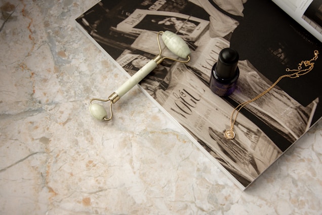 A facial roller, an essential oil bottle, and a gold necklace are placed on an open magazine with a monochrome print on a marble surface.