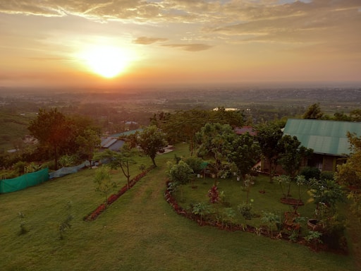 A serene landscape featuring a lush green garden with a variety of trees and plants. A small house with a green roof is nestled among the greenery. In the background, a panoramic view of a vast valley with distant hills is visible. The sun is setting on the horizon, casting a warm glow across the sky.