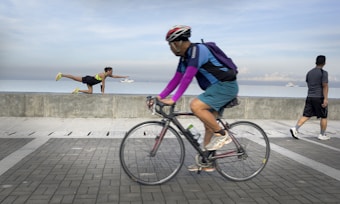 A cyclist wearing a helmet and bright sleeves rides along a paved path by the sea. A person in athletic clothing performs an impressive yoga or exercise pose on a concrete barrier, appearing to reach over a large ship in the distance. Another person walks leisurely in the same area, with the sea and cloudy sky creating a serene background.