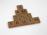 Wooden Scrabble tiles are arranged to form a pyramid shape with the words 'I AM THE TRUTH'. The tiles have black letters and numbers indicating their points value on a light wooden background.