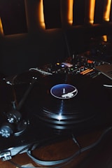A dimly lit scene featuring a turntable with a vinyl record spinning on it. The background consists of ambient lighting, creating a warm atmosphere. Nearby, there is an audio mixer with various controls and buttons, indicating a DJ setup.